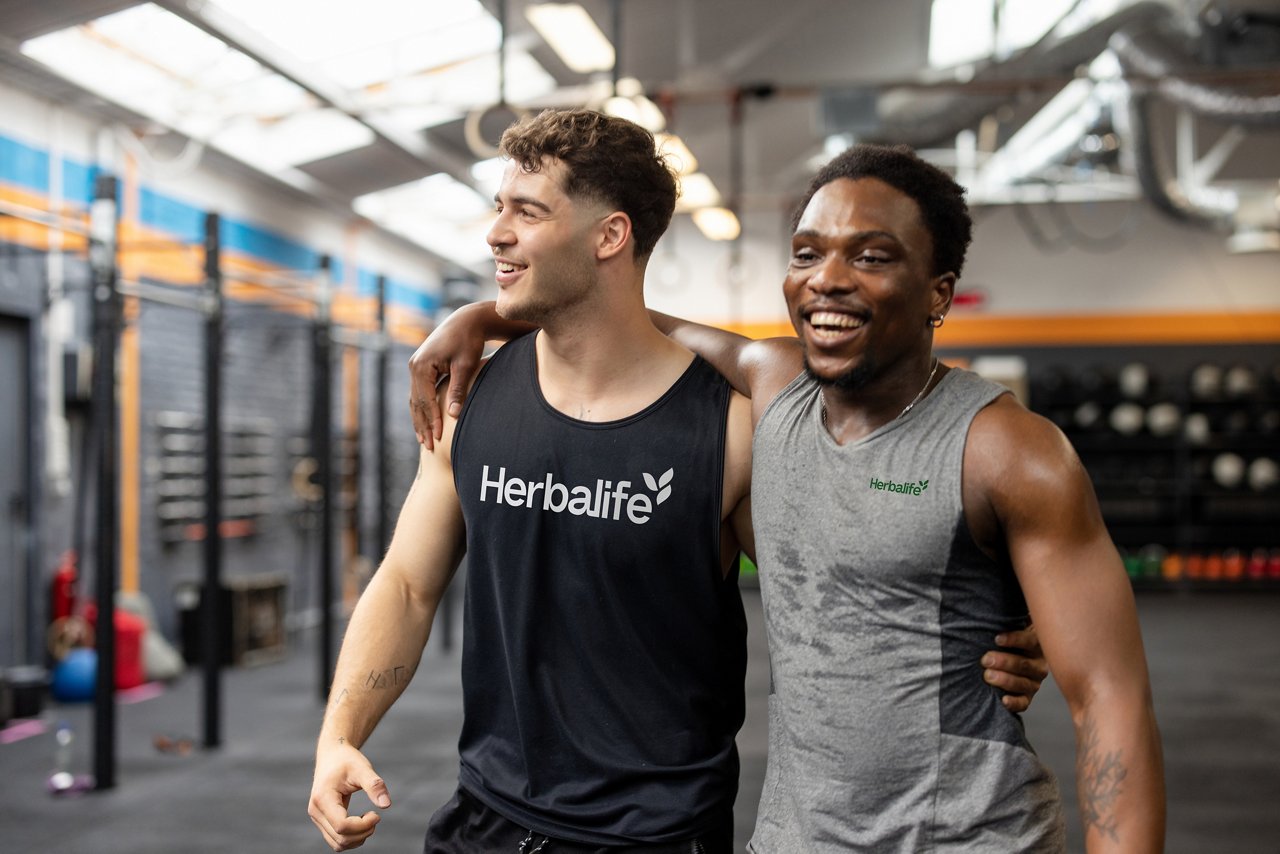 Two men at the gym laughing, one arm around the other, wearing herbalife workout clothes