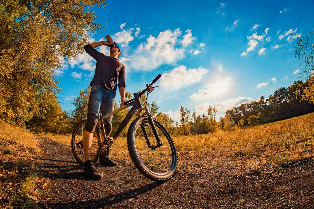 A young man drinks a protein shake from a shaker while taking a bike outdoors in the fall.
