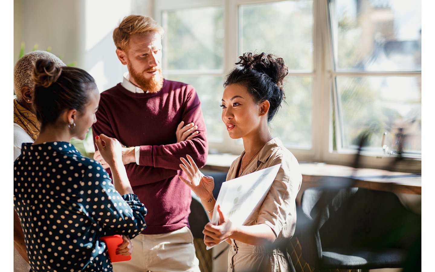 Colleagues standing in a small group discussing something. One of the women is holding documents and gesturing with her hands as the others watch and listen.