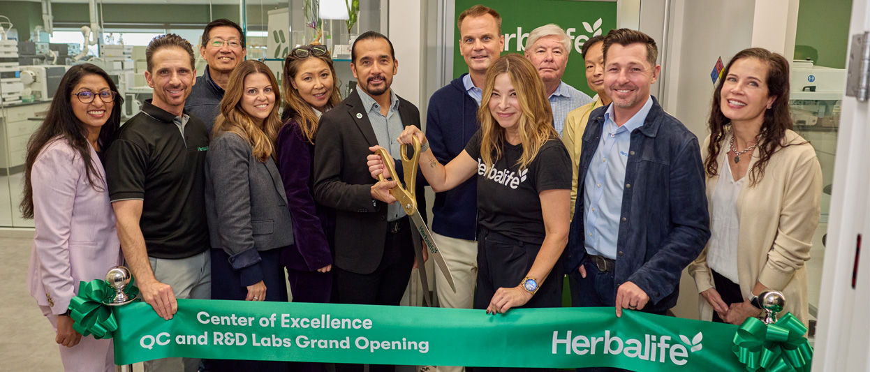 Ribbon-cutting ceremony for the Herbalife Center of Excellence QC and R&D Labs Grand Opening, featuring a green banner with Herbalife branding and participants holding oversized scissors inside a modern laboratory setting.