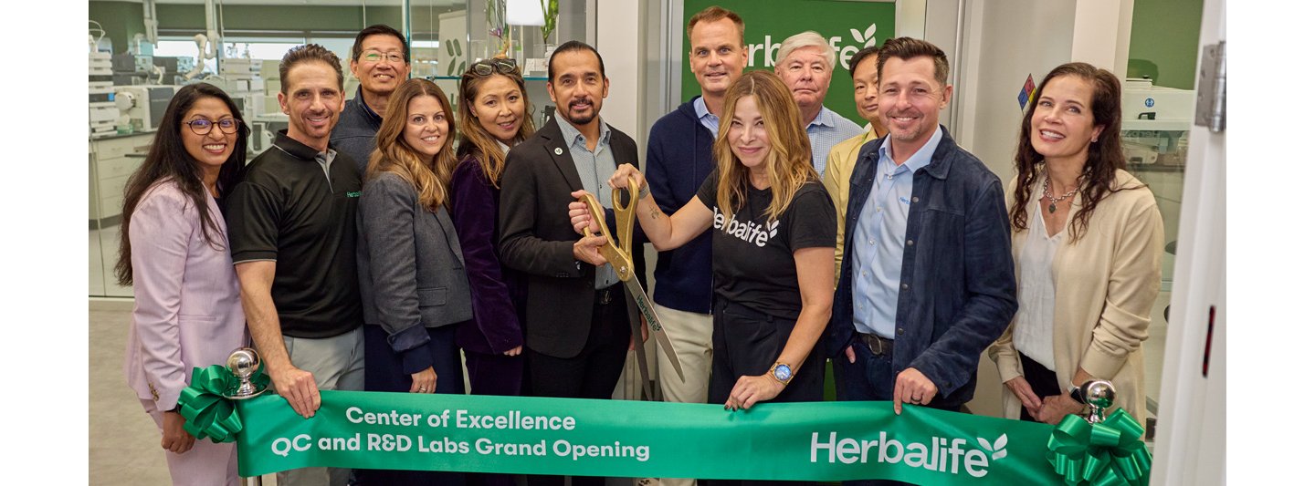 Ribbon-cutting ceremony for the Herbalife Center of Excellence QC and R&D Labs Grand Opening, featuring a green banner with Herbalife branding and participants holding oversized scissors inside a modern laboratory setting.