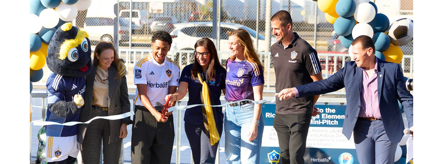 Ribbon-cutting ceremony at Ricardo Lizarraga Elementary School’s new mini-pitch in South Central Los Angeles, featuring LA Galaxy mascot, Herbalife representatives, and soccer-themed decorations with balloons and signage.